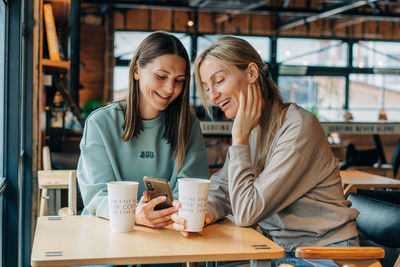 Two women sitting in a coffee house and laughing use the phone for online communication.