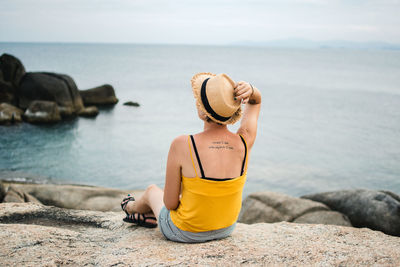 Rear view of man sitting on rock by sea against sky