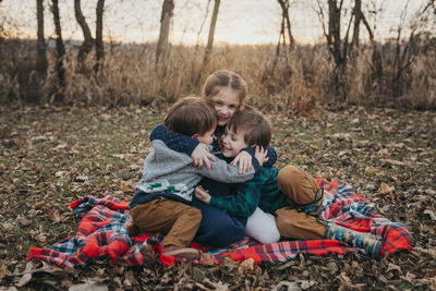 Three siblings sitting on red plaid blanket hugging at sunset