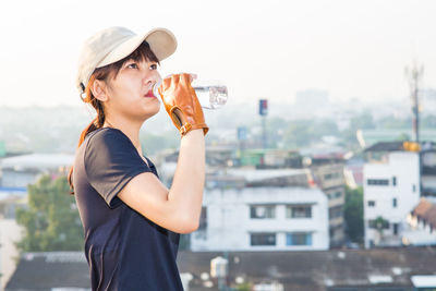 Young woman drinking water while standing by cityscape against clear sky