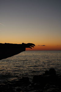 Silhouette hand by sea against sky during sunset