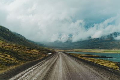 Empty dirt road leading towards mountains against cloudy sky