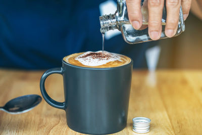 Close up of mature woman pouring warm syrup into hot cappuccino on cafe table. 