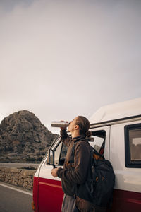 Young man drinking water while standing near van on vacation