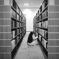 Woman reading book while sitting amidst bookshelves at library