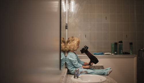 Side view of girl holding hair dryer while sitting by bathroom sink at home
