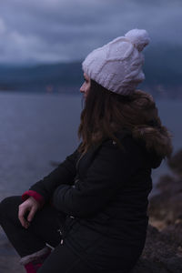 Woman looking away while sitting by lake against sky