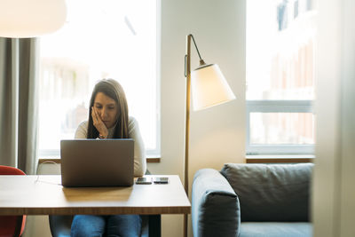 Thoughtful young hispanic female freelancer with long dark hair in casual clothes and earphones sitting at wooden table with hand on cheek and working remotely on laptop at home