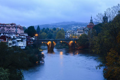 Arch bridge over river amidst buildings against sky at dusk