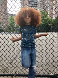 Portrait of boy standing against chainlink fence in city