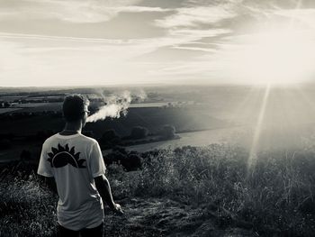 Rear view of man exhaling smoke while standing on hill looking at landscape against sky during sunset