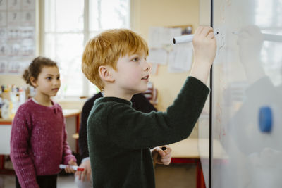 Side view of blond boy writing on whiteboard while solving mathematics in classroom