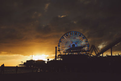 Silhouette santa monica pier by amusement park against cloudy sky during sunset