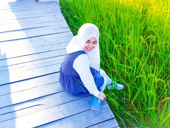 High angle view of smiling young woman wearing hijab while sitting on boardwalk at farm