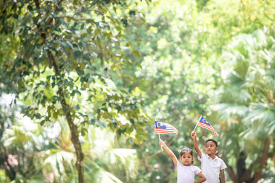 Portrait of siblings holding malaysian flag while standing against trees at park