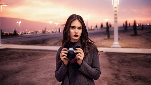 Portrait of young woman photographing against sky during sunset