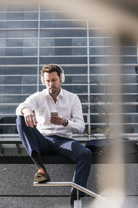 Businessman sitting in front of office building, wearing headphones