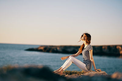 Woman sitting by sea against clear sky during sunset