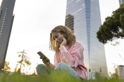 Smiling young woman using mobile phone on grass in city