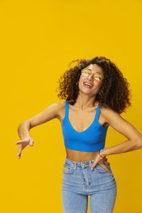 Portrait of young woman with arms raised against yellow background