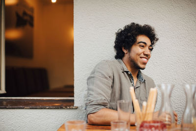 Smiling young man looking away while sitting at table against wall