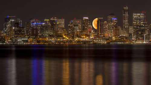 Illuminated buildings by river against sky at night