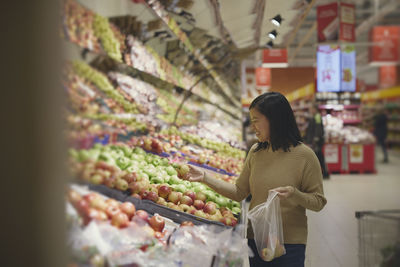 View of woman standing in supermarkets and putting apples into plastic bag