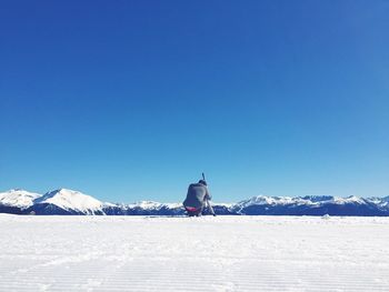 Rear view of man crouching on snowy field against clear blue sky