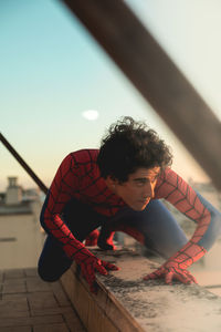 Young man in spider-man costume kneeling on building terrace against sky