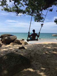 Rear view of man sitting on rock at beach against sky