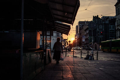 Rear view full length of woman walking on sidewalk during sunset in city