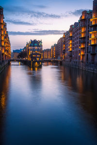 Illuminated buildings by river against sky at sunset