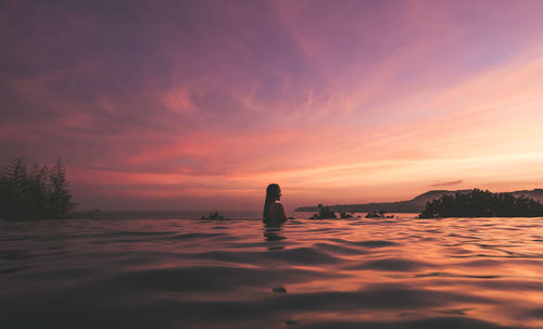 Young woman swimming in infinity pool by sea against sky during sunset