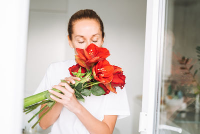 Young woman in white t-shirt with bouquet of red flowers in hands near window in kitchen at home