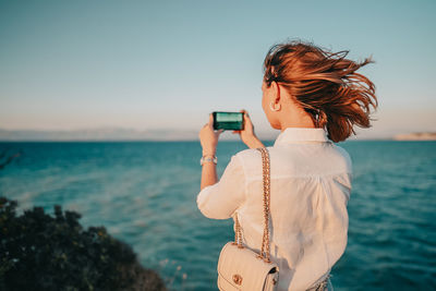 Woman photographing while using smart phone against sea