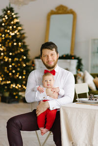 A stylish dad in a white shirt with a red bow tie holds his son in his arms against the background