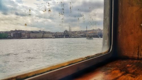 Buildings by sea against sky seen through glass window