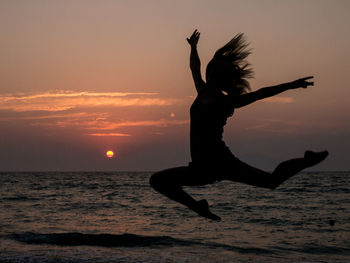 Silhouette woman with hand raised jumping at beach against sky during sunset