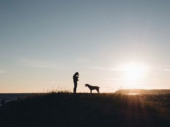 Silhouette man with dog on field against sky during sunset