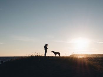 Silhouette woman with dog on field against sky during sunset