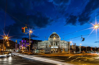 Light trails on city street by building against sky at dusk