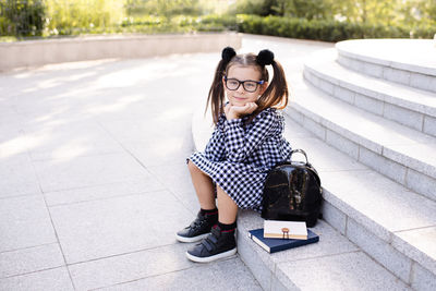 Smiling happy kid girl 5-6 year old wear uniform dress and glasses sitting on stair with books