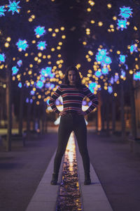 Portrait of woman standing on footpath against illuminated lights at night