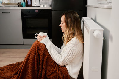 Worried woman sit near heating radiator under blanket with cup of tea