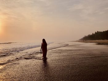 Rear view of woman walking at beach against sky during sunset