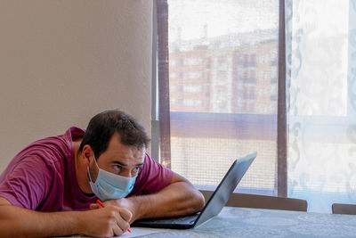 Side view of man wearing flu mask using laptop at home