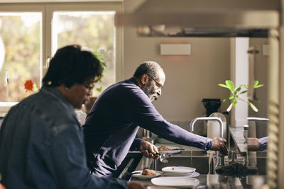 Senior man turning on faucet while washing spoons in kitchen at home