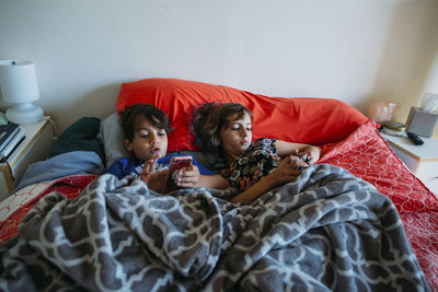 High angle view of siblings using smart phones while lying on bed