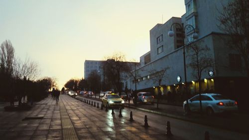 Cars on road by buildings against sky during sunset