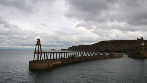 Beacon on pier in sea against cloudy sky